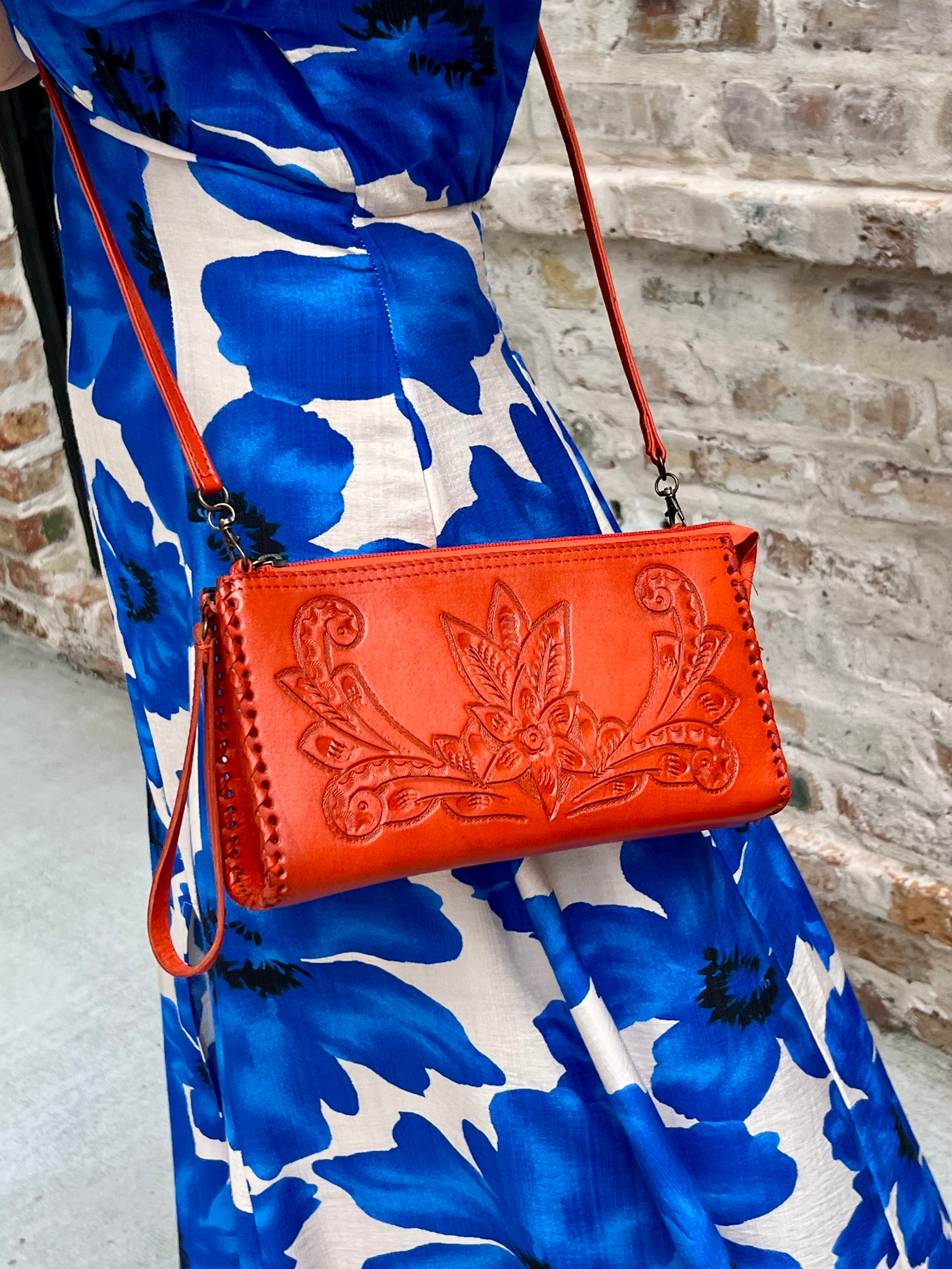 Red leather handbag with floral embossing on a blue floral dress against a brick wall.