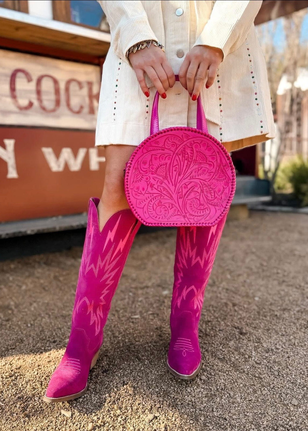 Person wearing pink cowboy boots and holding a pink handbag in front of a wooden sign.